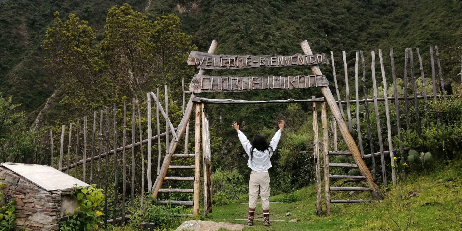 Turista celebra al llegar a Choquequirao. Arco de madera con letrero "Welcome - Bienvenido Choquequirao" en medio de montañas verdes