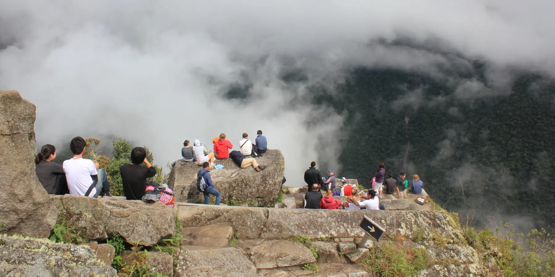 Visitantes sentados sobre rocas en un mirador alto de Huayna Picchu, contemplando el valle cubierto de nubes y neblina sobre el bosque del Urubamba.