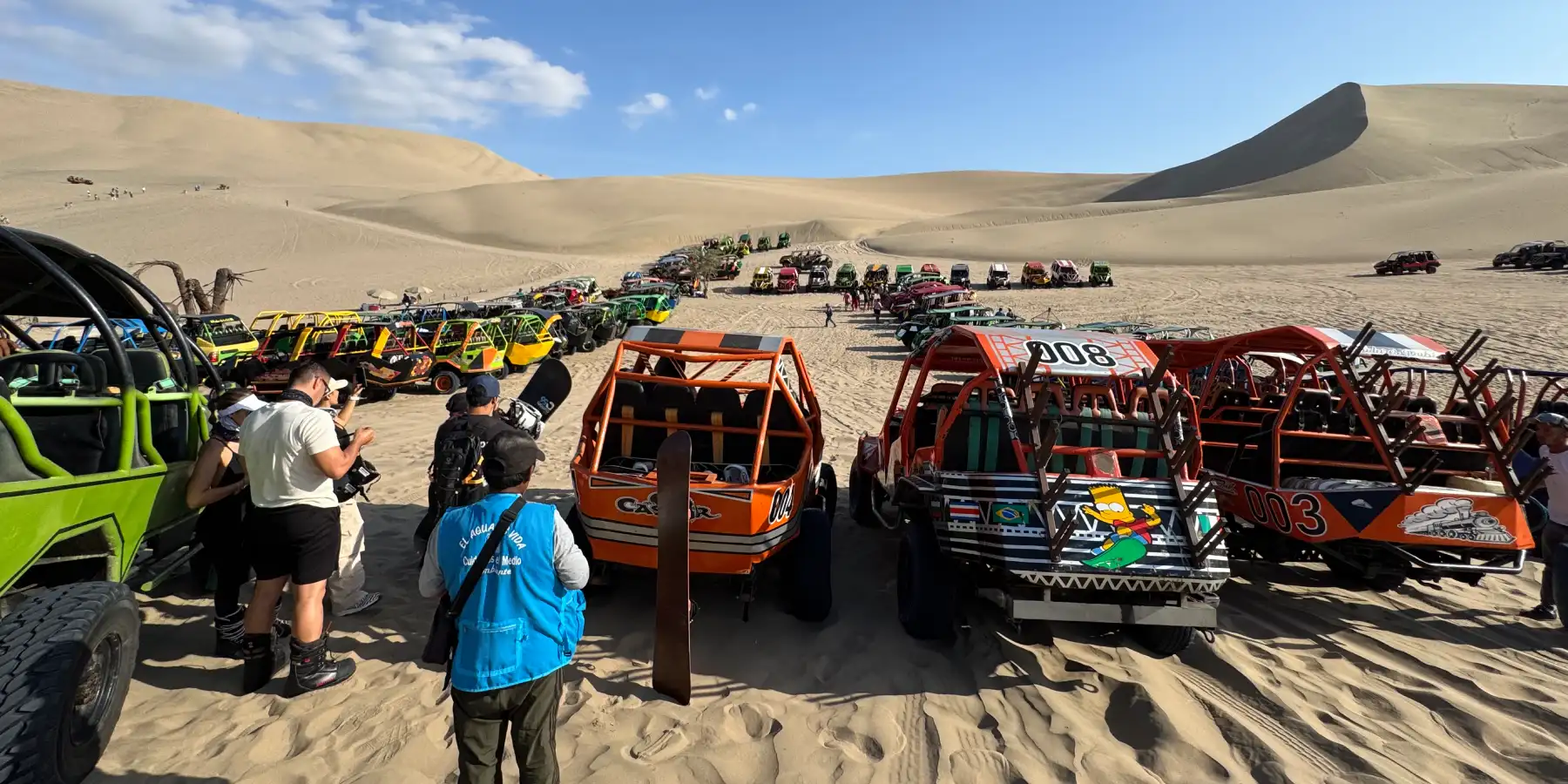 Gran punto de encuentro en las dunas de Huacachina (Ica, Perú), con decenas de buggies alineados y turistas con tablas/equipo de sandboarding antes de iniciar el recorrido.